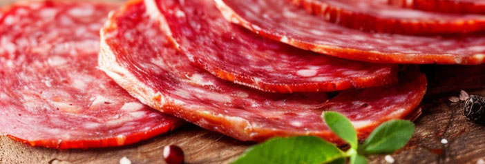 Thin slices of organic Genoa salami fanned out on a wooden board with fresh herbs and peppercorns